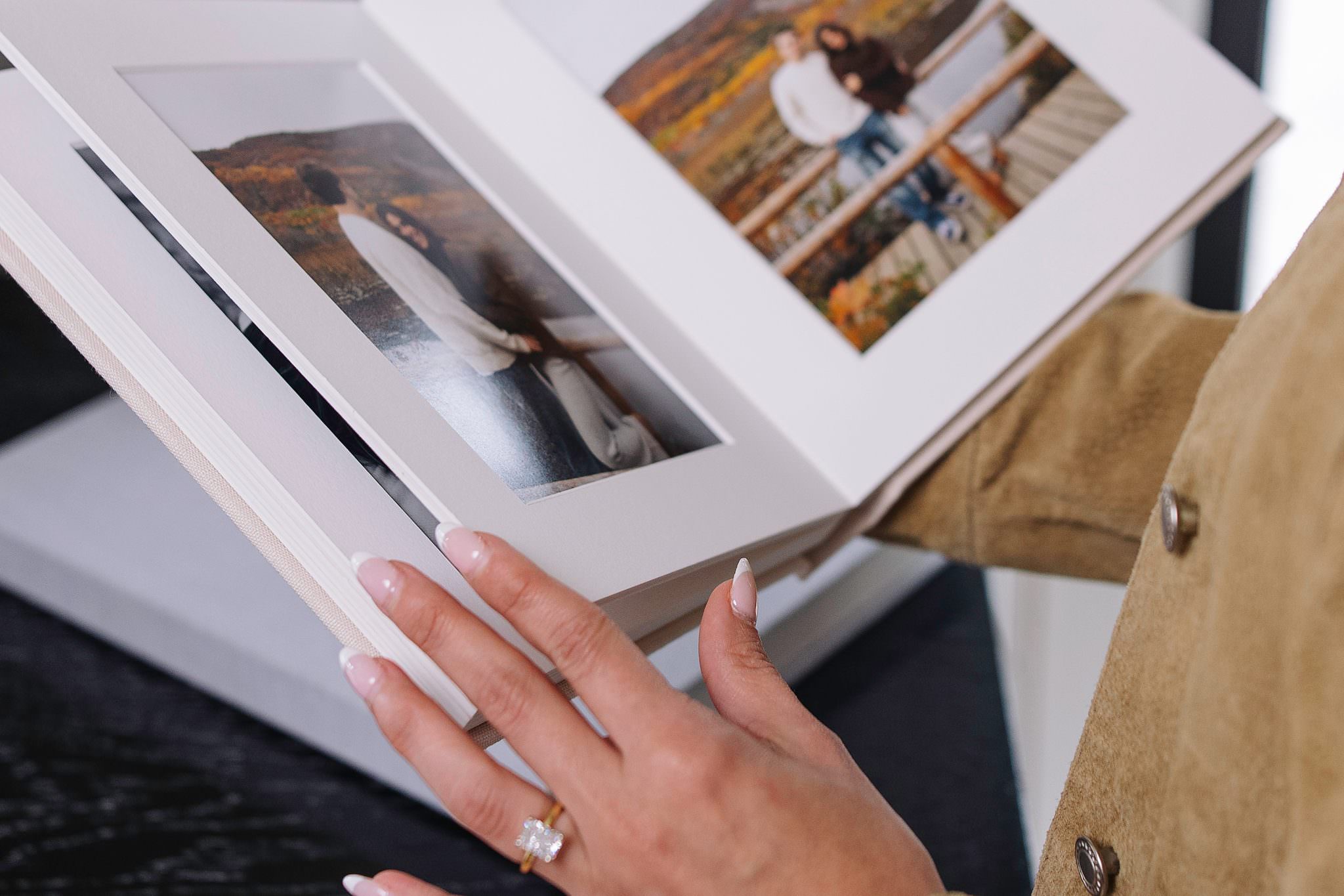 An open matted folio album showing two side by side family portraits in white mats, held by a hand with an engagement ring