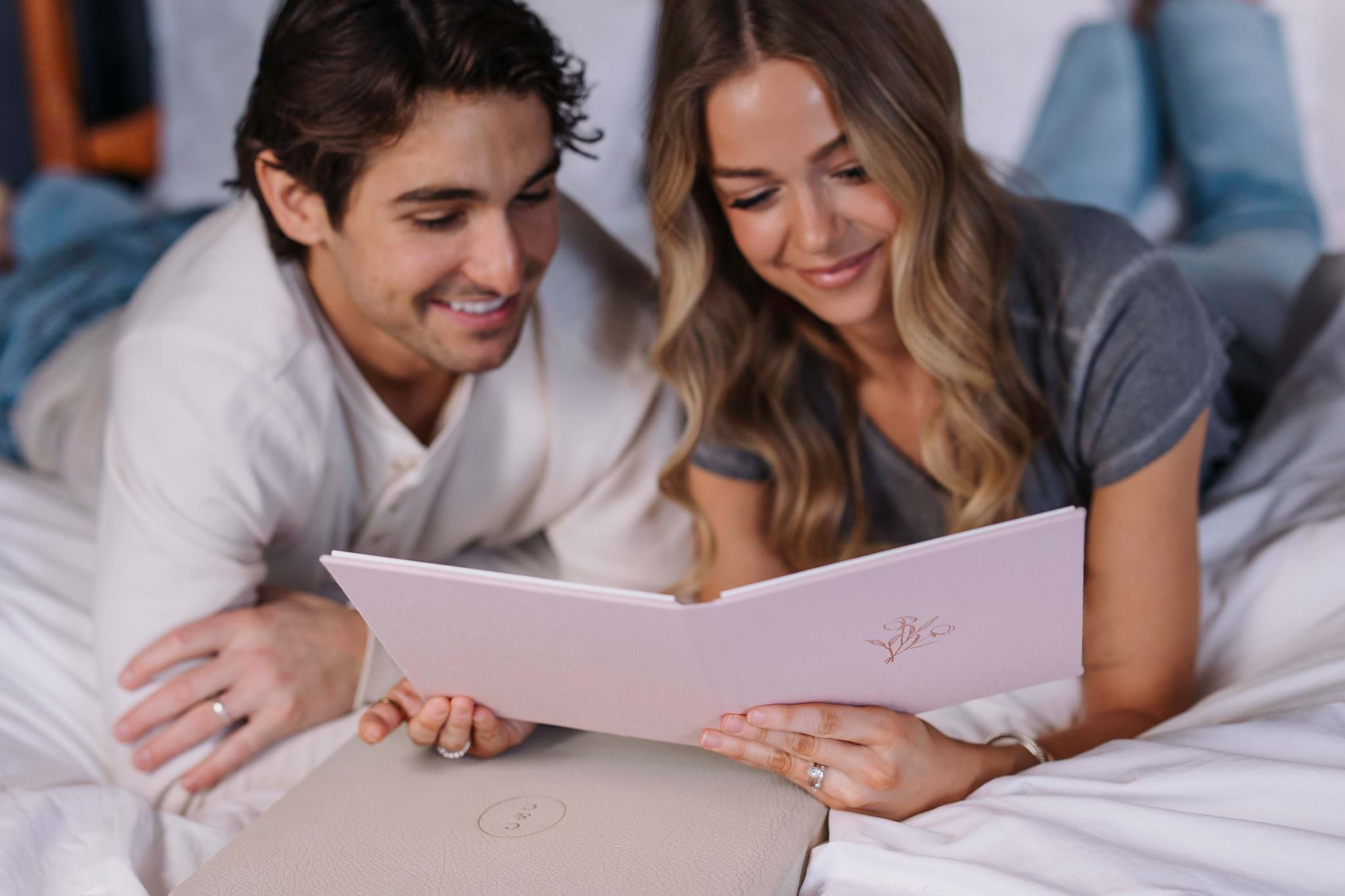 A couple lying on a bed opening a blush matted folio with a rose gold debossed monogram