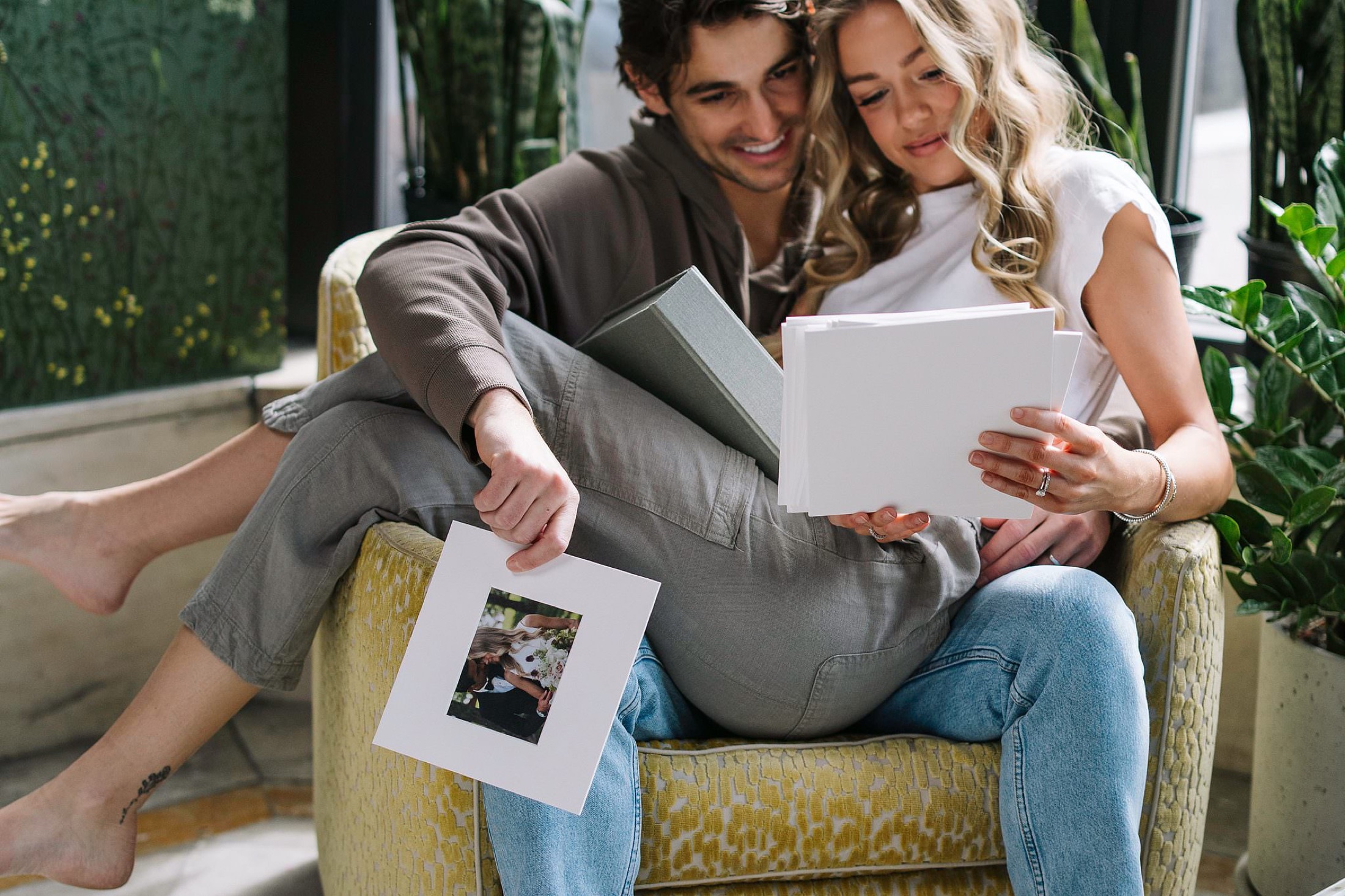 A couple together in a chair, leafing through a stack of matted prints in afternoon light